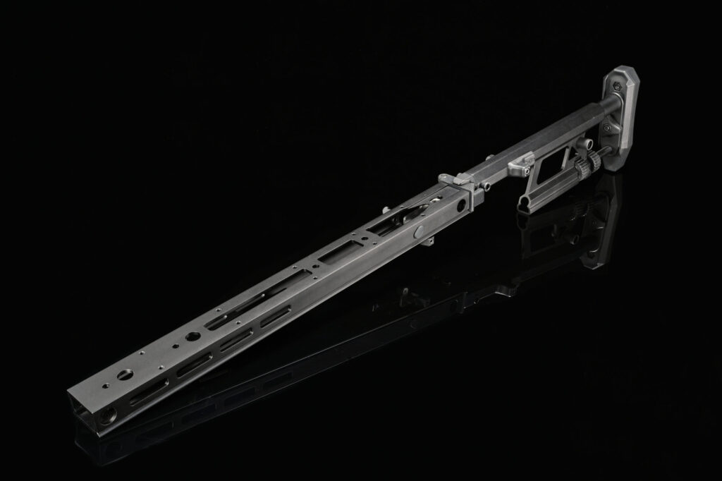 This image displays another angle of the tactical firearm you previously showed, specifically focusing on its top side. The elongated handguard with multiple cutouts and ventilation holes is prominent in this perspective. The adjustable stock is also visible, and from this angle, one can see the intricate details and design of the buttstock. The firearm retains its matte gray finish, and the reflection below adds to the dramatic presentation against the dark background.





